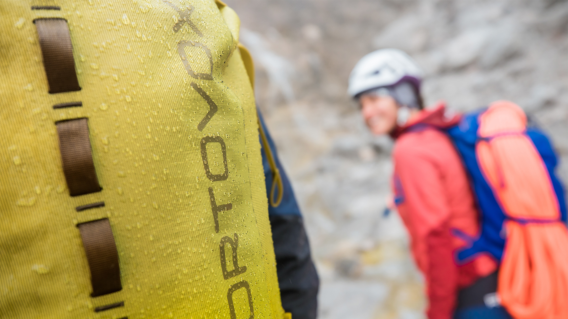 黄色いORTOVOX DRYバックパックの水滴と、背景でぼけて見える2人の登山者