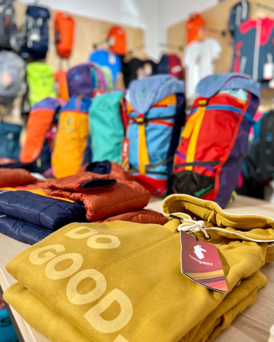 店内で畳まれたCotopaxi Do Goodのフーディーと背景の色鮮やかなバックパック