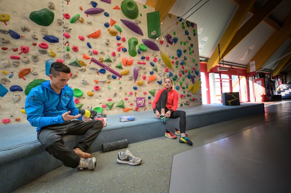 クライミングジムの色とりどりのホールドが付いた壁の前に座る二人 — Jessica Pilz of SCARPA, credits Tristan Hobson