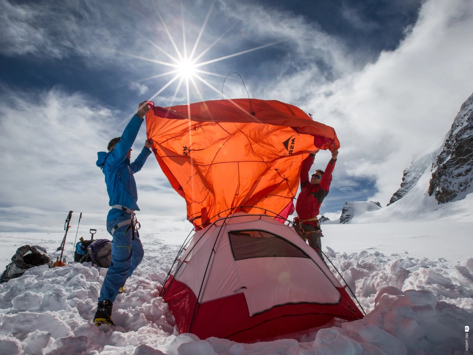 雪の上のテントの上でオレンジ色のシートを持ち上げる二人 — Credits to Eric Larsen, Msr Gear