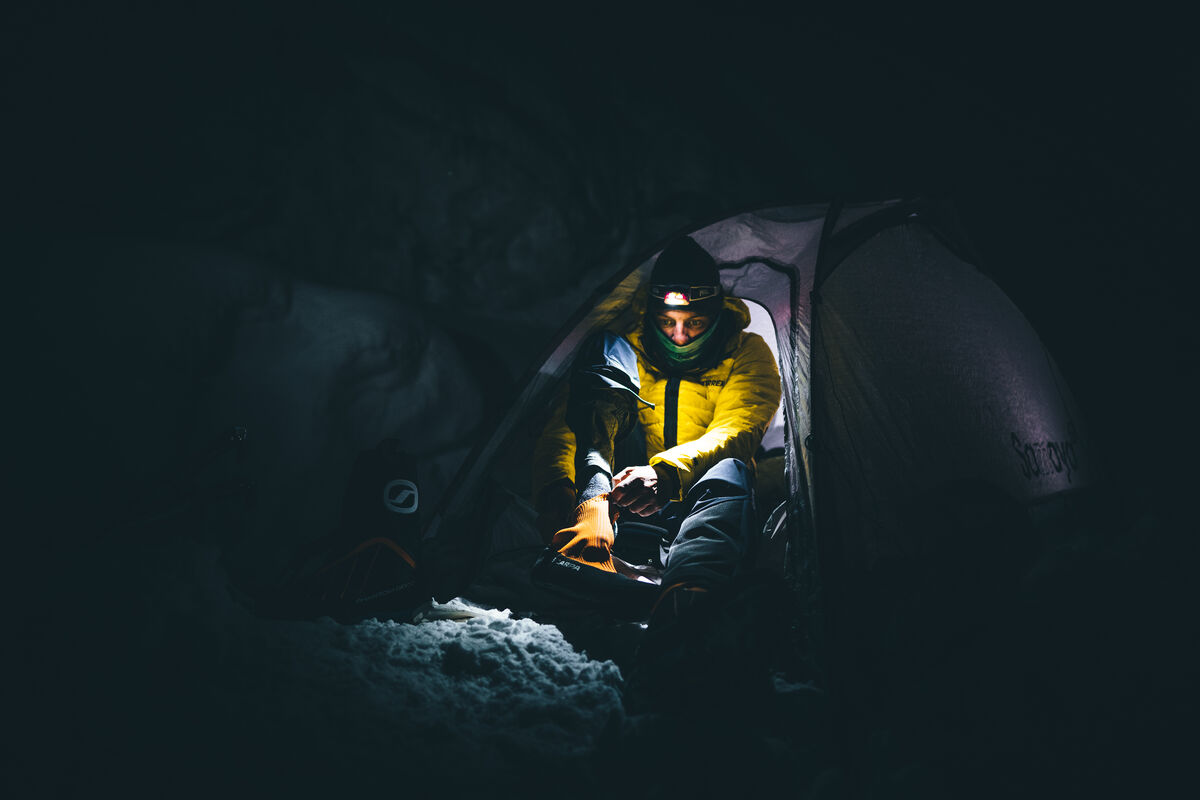 暗闇の中、雪の上のテントの入口にヘッドランプを着けて座る人 — ©Mathis Dumas, athlete Mathieu Maynadier_location Mont Blanc area _