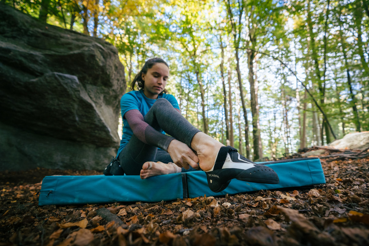 森の中の青いクラッシュパッドに座ってクライミングシューズを履くクライマー。写真 Antoine Mesnage / SCARPA