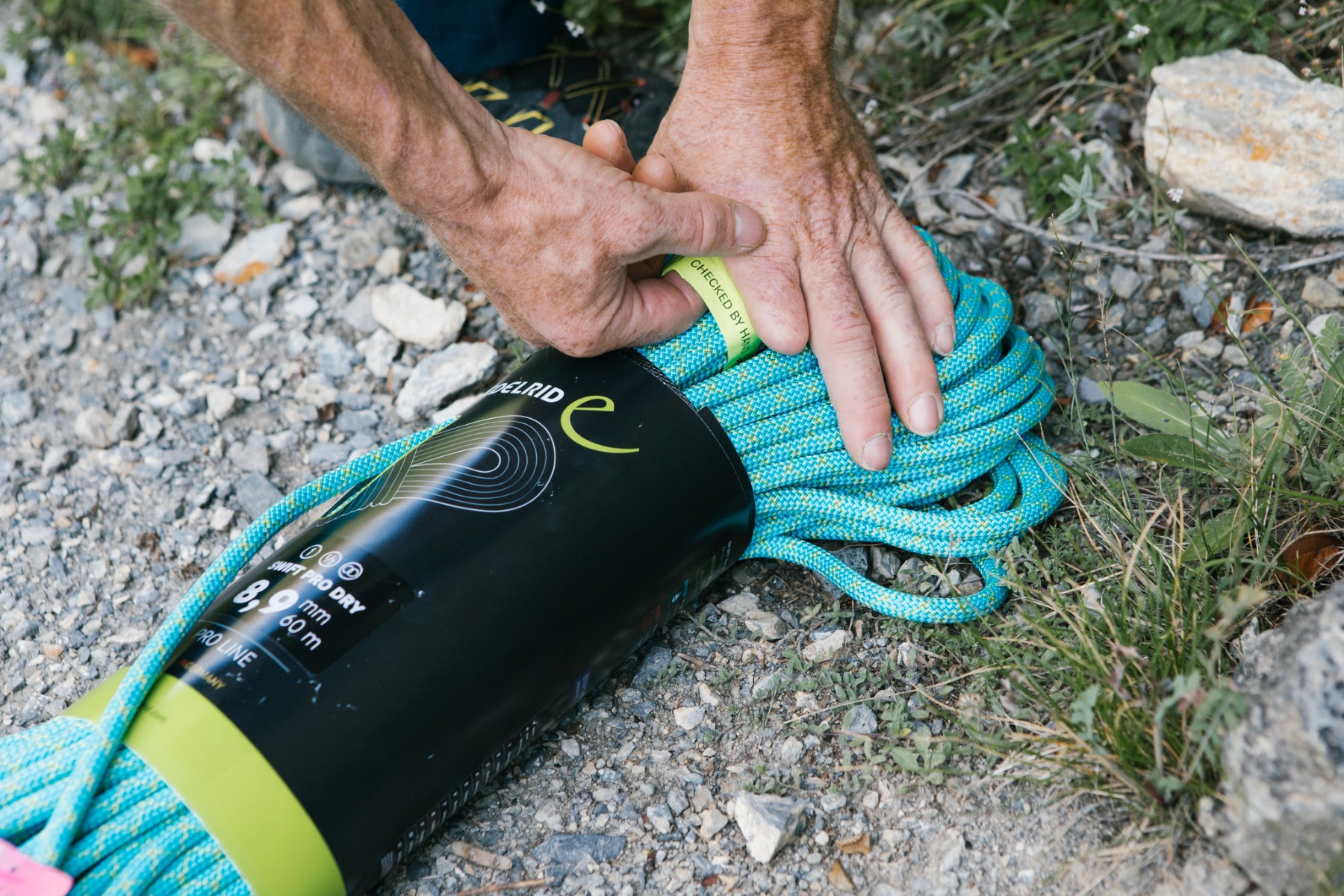 石の多い地面で、Edelrid SWIFT PRO DRY 8,9 mmのパッケージに入った青いロープの束を整える手