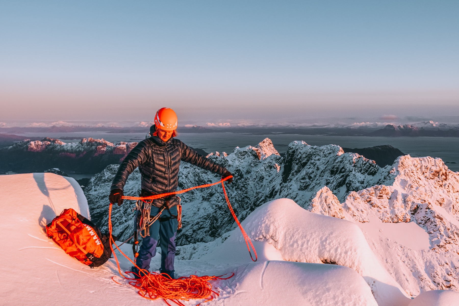 雪の稜線で、ヘルメットとハーネスを着けてオレンジ色のロープを扱う人。背景には山々が見える
