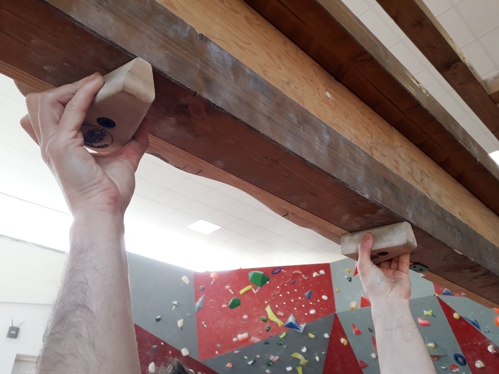 Pinze Owl Climb, 自宅でのトレーニング 木の梁に取り付けられた2つの木製ホールドを握る両手。背景にはクライミングウォールが見える