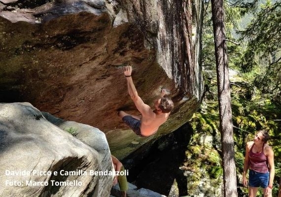 davide picco bouldering magic wood 上半身裸で暗い色のズボンを着た人が、傾斜の強い岩にいる。その隣には、紫色のトップスと青いショートパンツを着た人が木々の間に立っている。