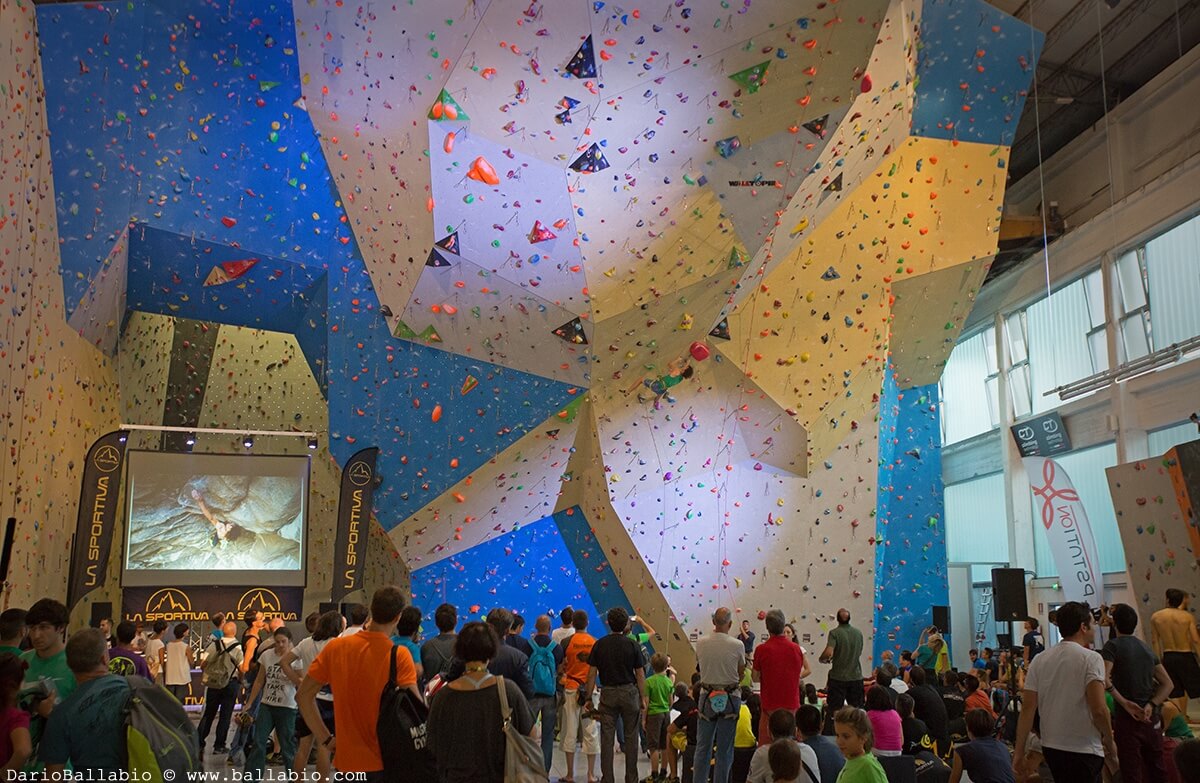 Milano Climbing Expo カラフルなホールドのある屋内クライミング壁。その前に観客が集まり、横にスクリーンがある