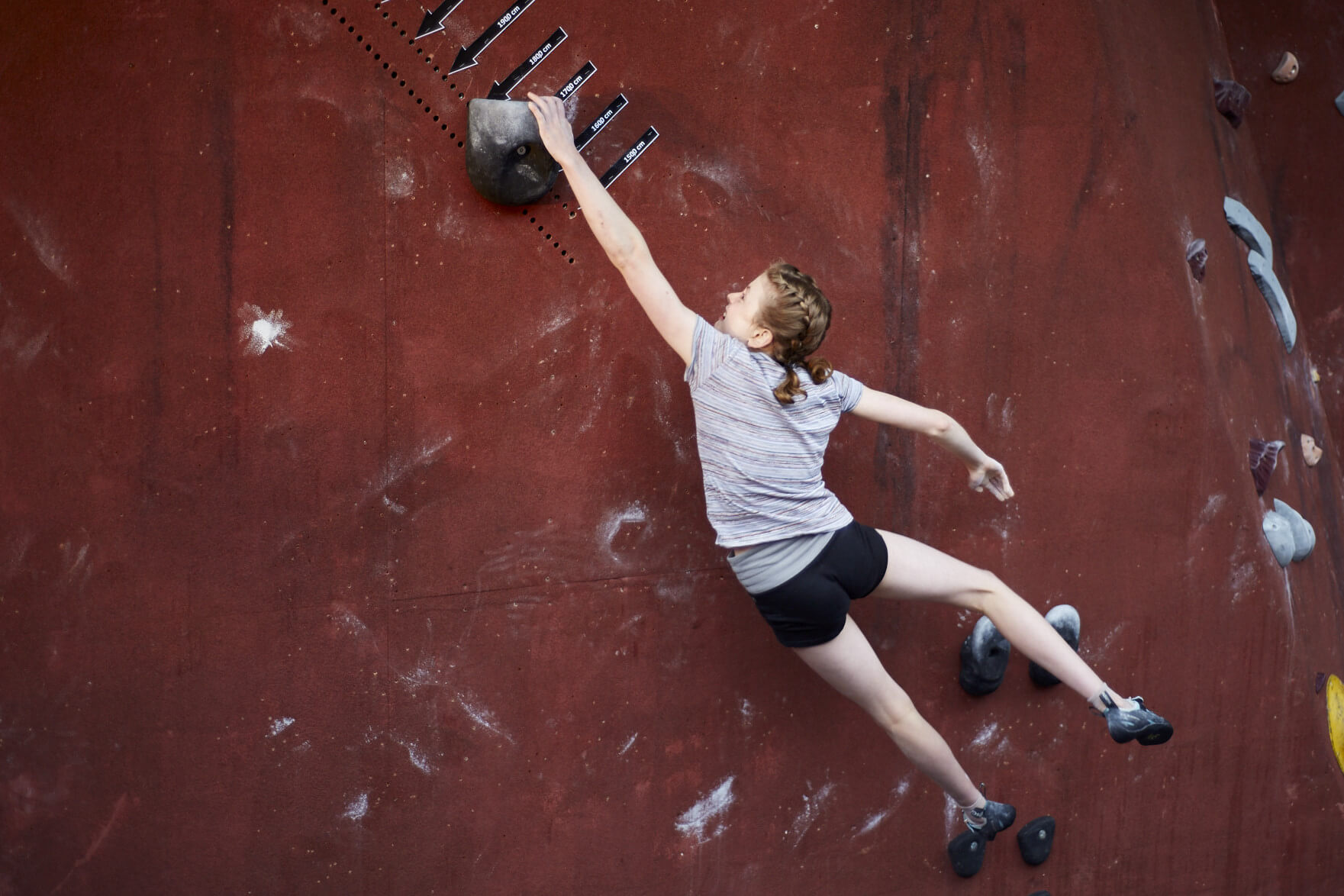 Gara di lanci, credits: the castle climbing 縞模様のシャツと短パンを着た人が、赤い屋内クライミング壁のホールドに手を伸ばしている, credits: the castle climbing