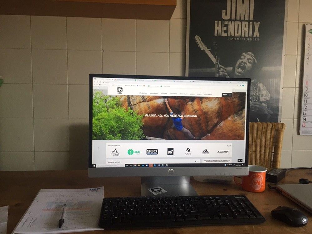 モニターにOliunìdのサイトが表示された作業スペース。キーボード、マウス、カップが置かれている