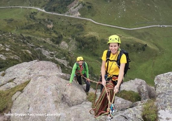 山の岩場でヘルメット、ロープ、ハーネスを着けた2人。1人は黄色いシャツとバックパック、もう1人は緑のジャケット