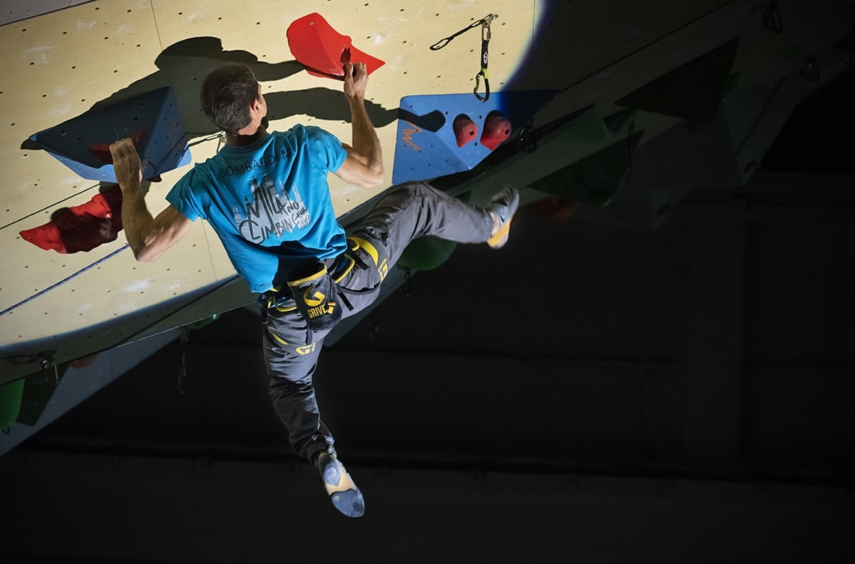 Milano Climbing Expo 赤いホールドと青いボリュームがある屋内の前傾壁で登る、青いTシャツとハーネスを着けた人