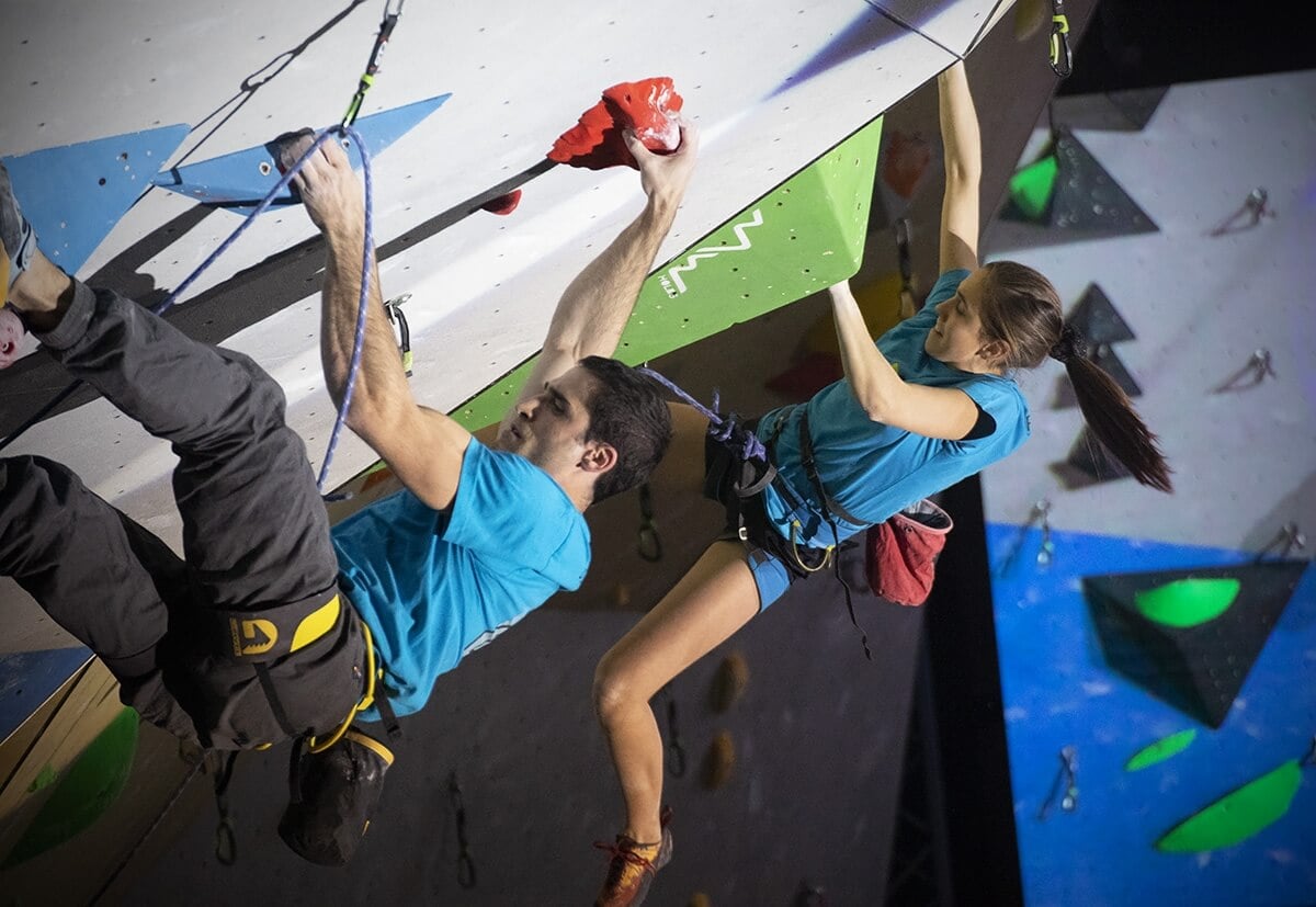Milano Climbing Expo 青いTシャツを着た2人が、ロープとハーネスを使って、色とりどりのホールドがある屋内の壁を登っている