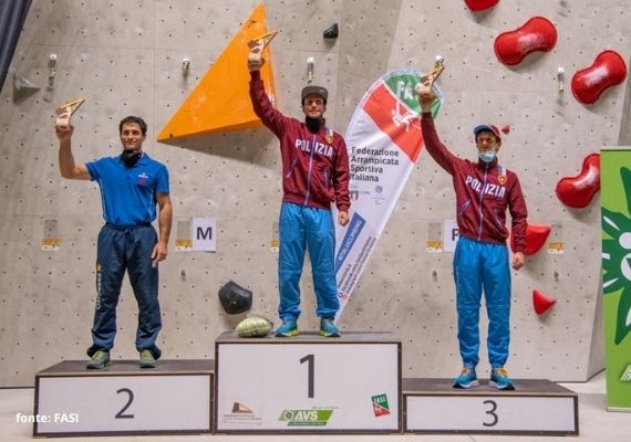 2020年イタリア・リードクライミング選手権の男子表彰台 屋内クライミング壁の前にある番号付きの表彰台で、トロフィーを掲げる3人。2人は赤い上着、1人は青いシャツを着用